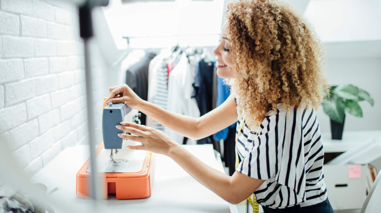 Female clothing designer using sewing machine next to rack of clothes.
